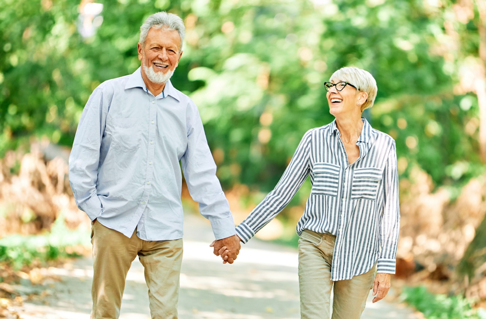 Smiling senior couple holding hands while walking outdoors on a sunny day, enjoying light physical activity and connection in a natural park setting.