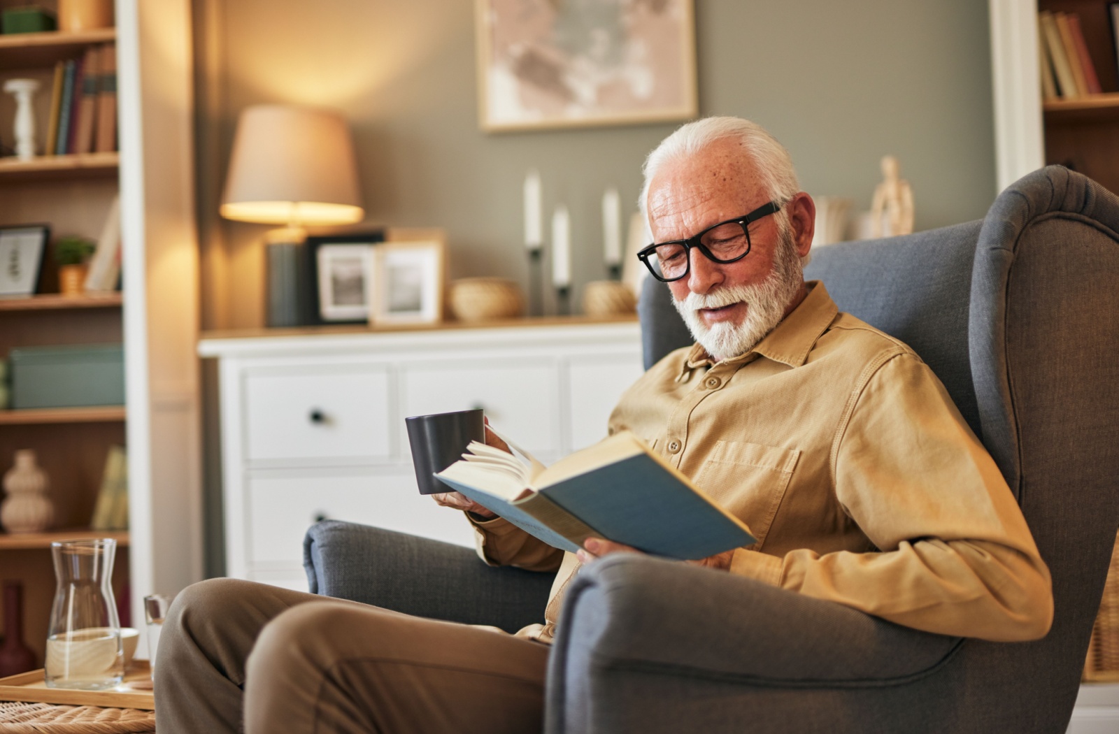 An older adult smiles while reading a hardcover book over coffee while sitting in their armchair in a living room