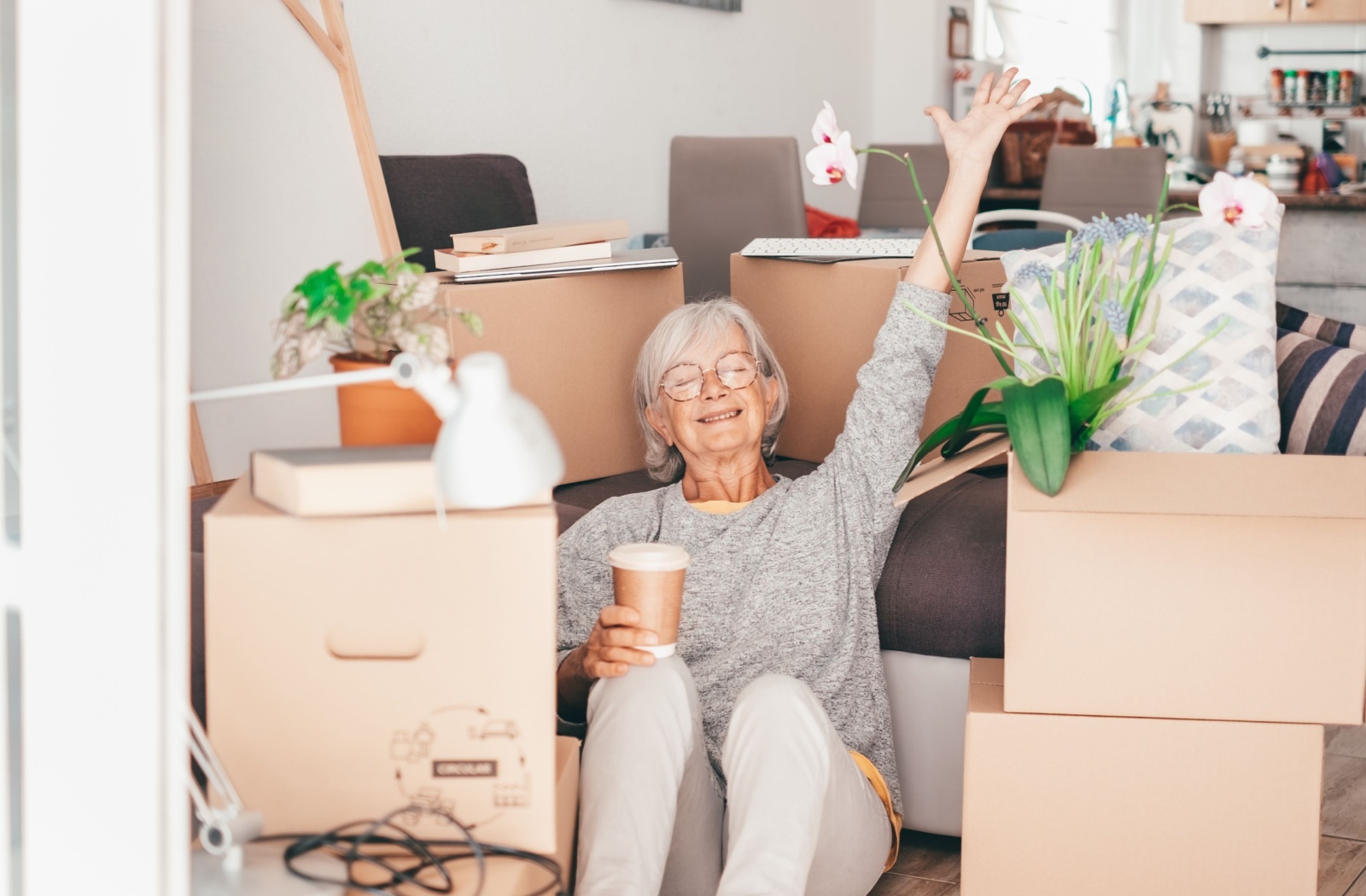 An elderly woman who is packing to move into senior living sits among packed boxes and smiles while holding a coffee