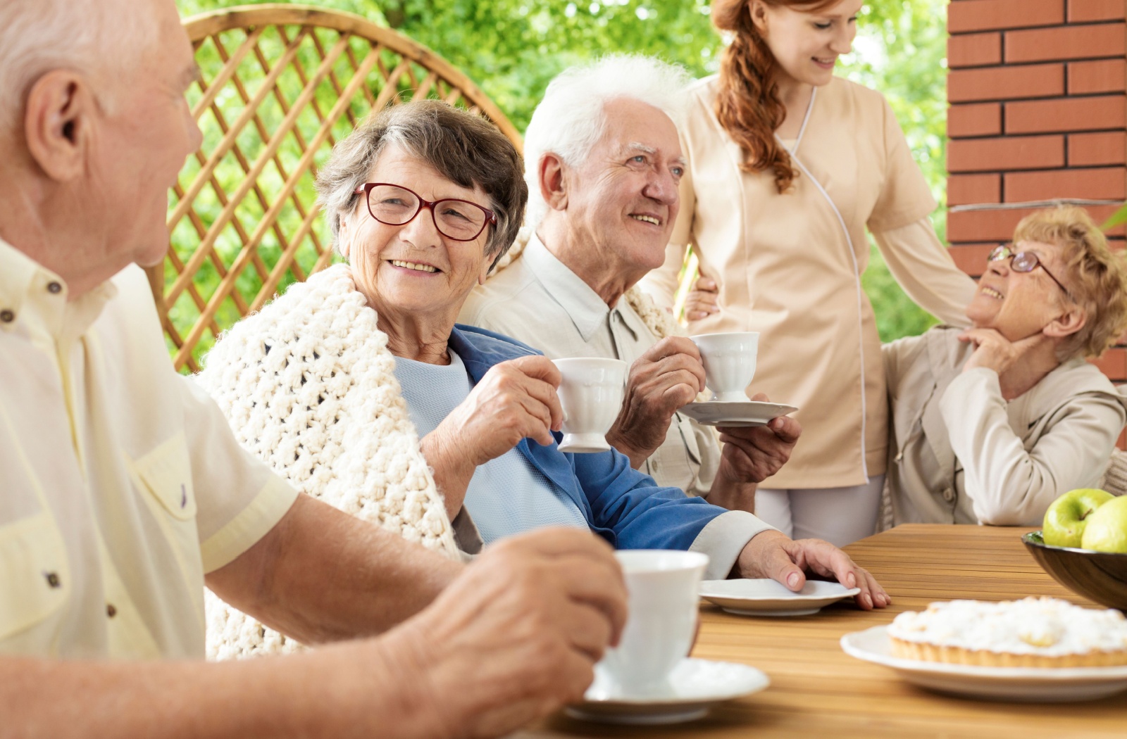 A group of seniors laugh and talk over tea at a table in their senior living community while an attendant watches them