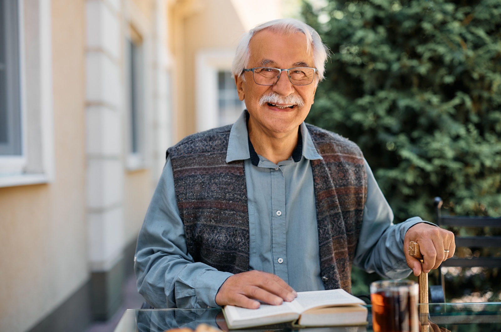 A smiling independent older adult sitting outside on a patio and reading a book.