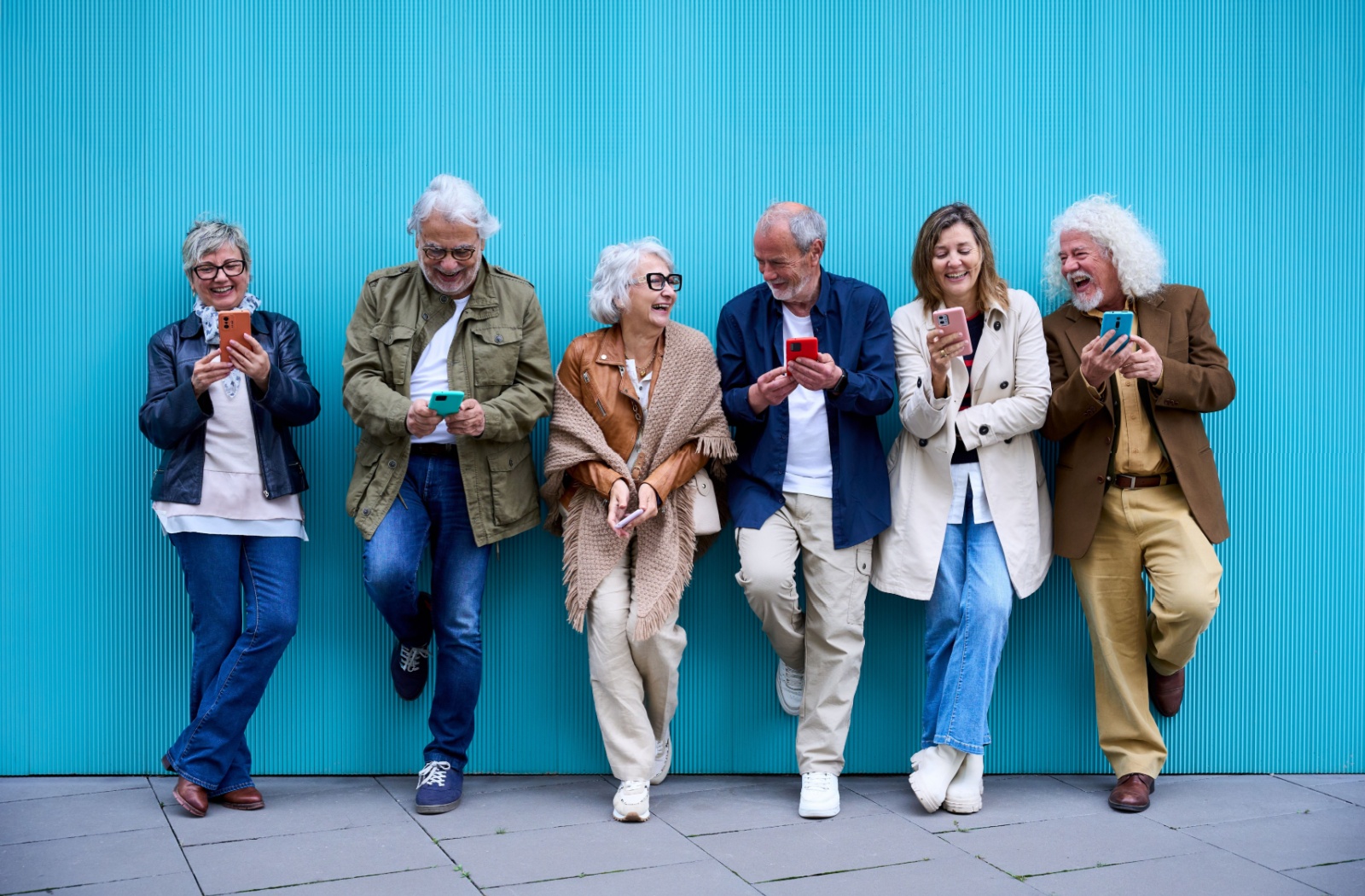 Group of seniors laughing together using their cell phones standing leaning on blue wall.