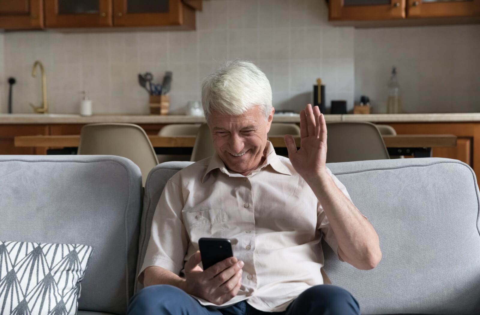 A smiling older adult sitting on the couch and waving while on a video call.
