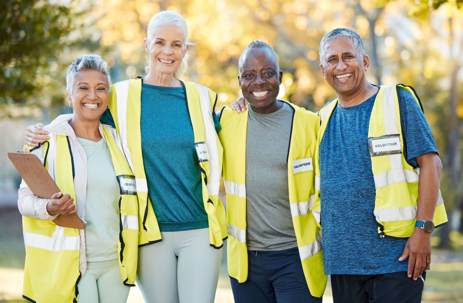 A group of smiling retirees in neon vests standing arm-in-arm in a park while volunteering together.