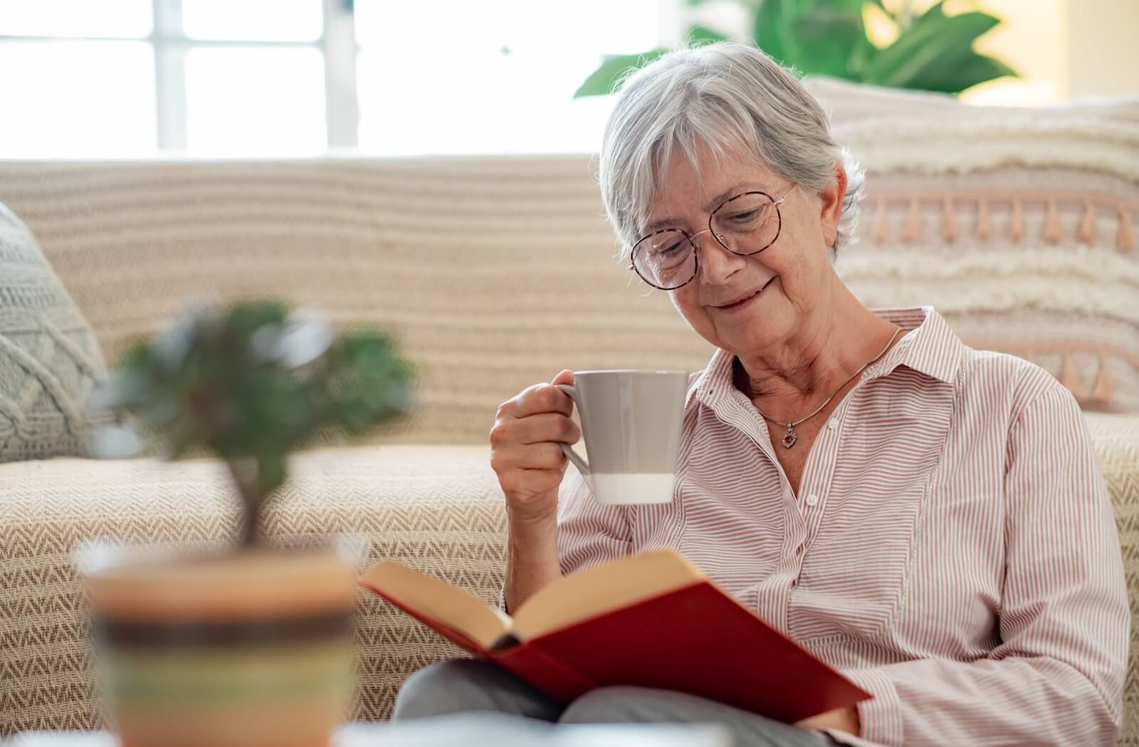An older adult smiles and takes a sip of their tea while reading a novel on their couch in senior living