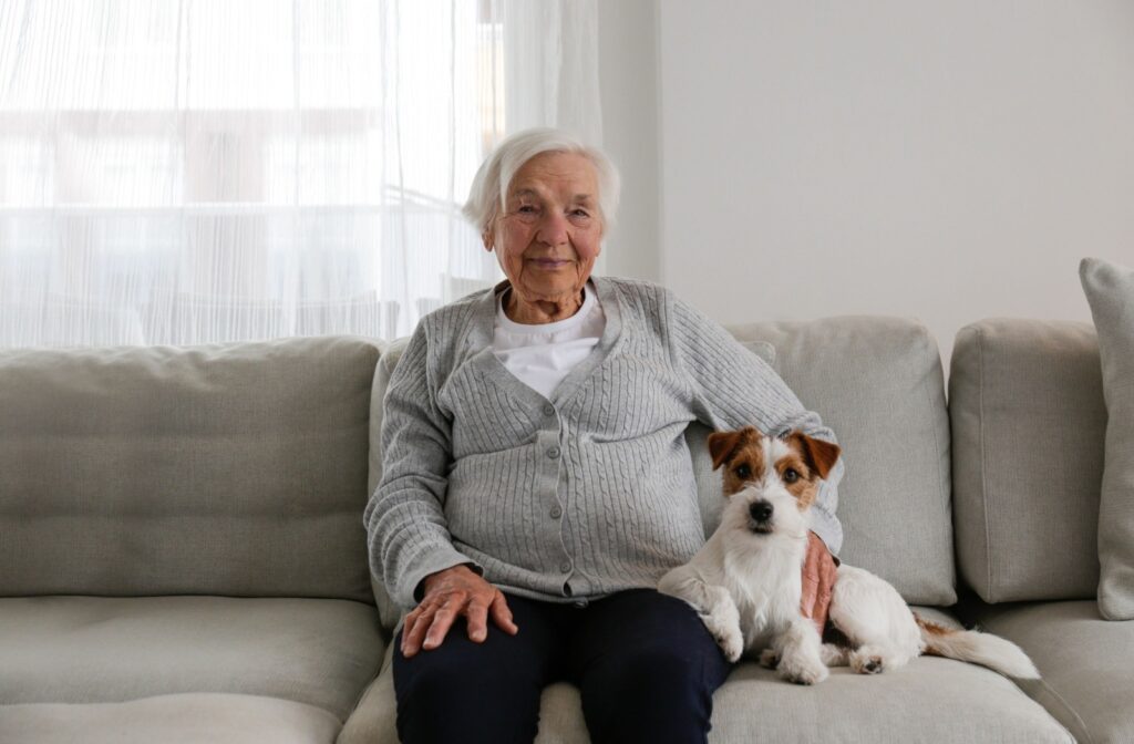 A senior resident sits on a gray couch next to their pet terrier.